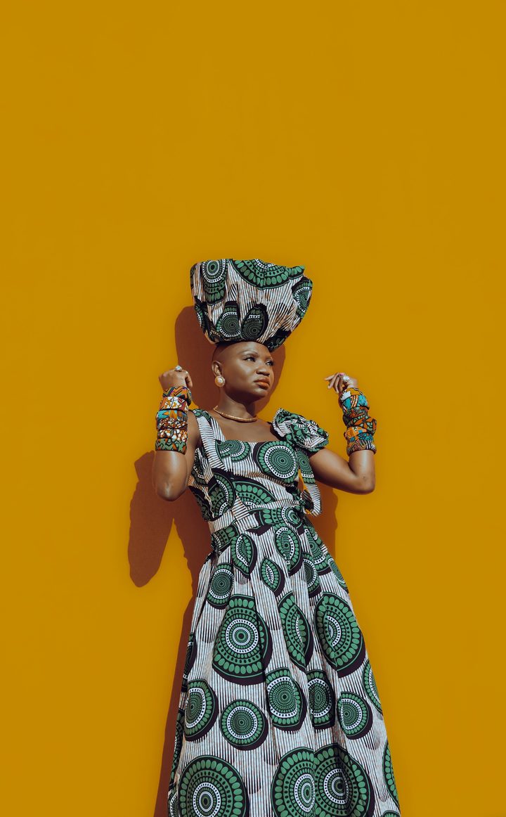 Femme posant devant un mur jaune dans une robe longue en wax vert et blanc, coiffée d'un turban assorti