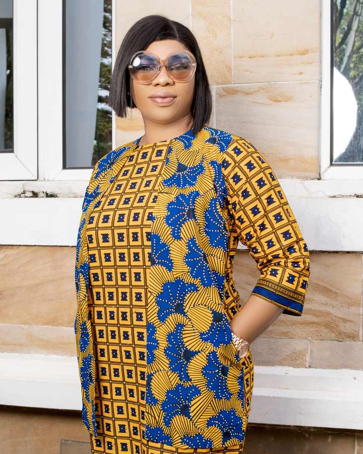Femme à lunettes de soleil vêtue d'une robe tunique en wax jaune et bleu, devant un mur en pierre