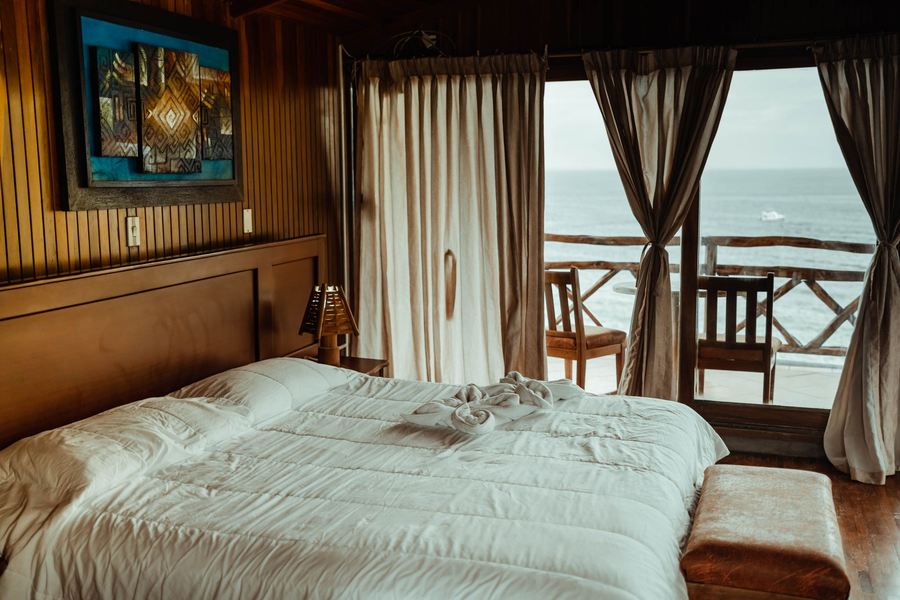 Chambre boisée avec grand lit blanc et rideaux ouverts sur un balcon face à la mer