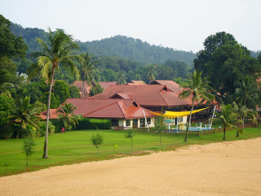 Bâtiments aux toits rouges entourés de cocotiers et de pelouse, au pied de collines boisées