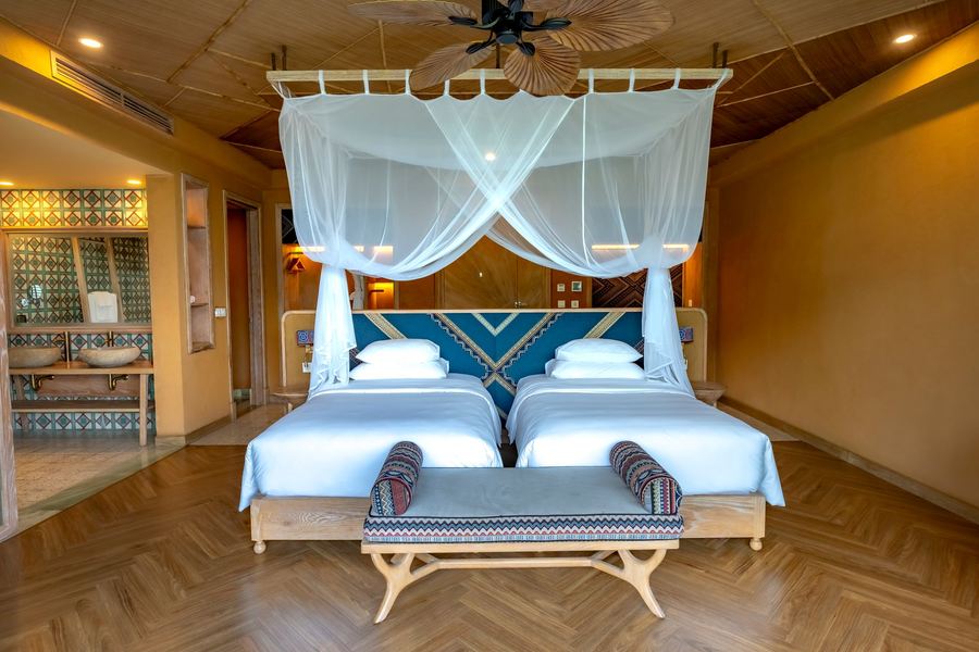 Grande chambre claire avec deux lits à baldaquin, tête de lit bleue et banc tissé au pied des lits